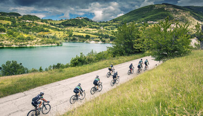 La foto mostra un panorama collinare, con un lago sulla sinistra, e dal basso arrivano dei turisti in bicicletta.
