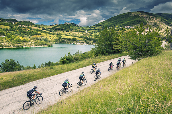 La foto mostra un panorama collinare, con un lago sulla sinistra, e dal basso arrivano dei turisti in bicicletta.