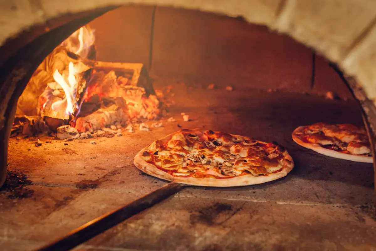 L'immagine mostra un forno a legna per la pizza, acceso, con una pala che sta tirando fuori la pizza cotta.